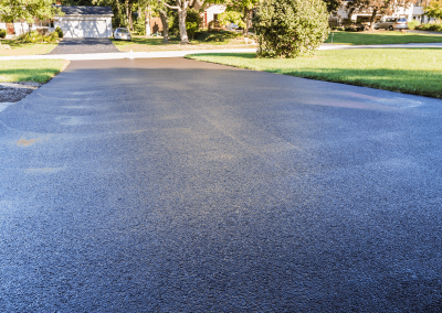 Freshly paved black asphalt driveway in a suburban neighborhood with green lawns and trees.