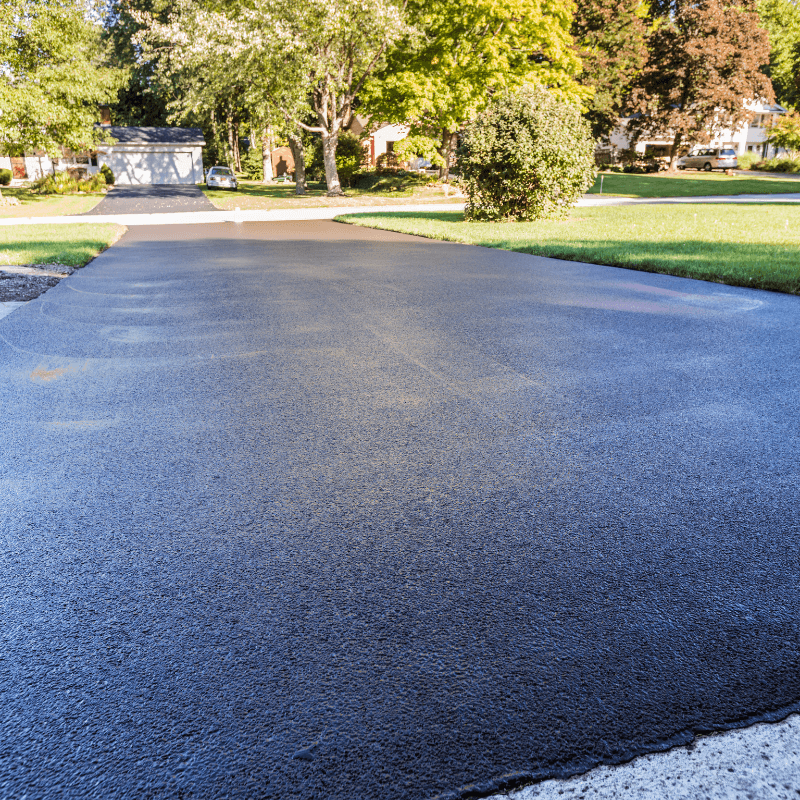 Freshly paved black asphalt driveway in a suburban neighborhood with green lawns and trees.