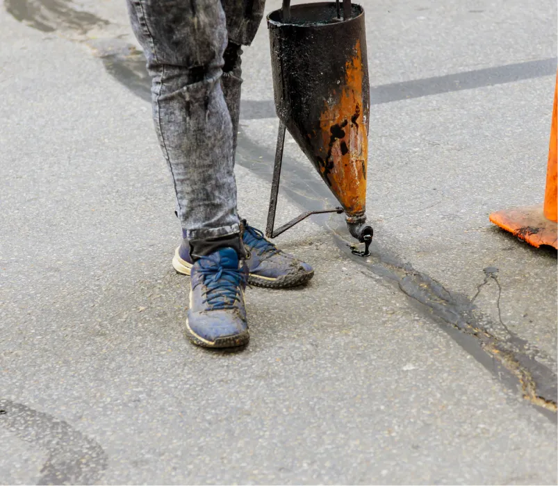 Person sealing a road crack with tar next to an orange traffic cone, only legs and tool visible.