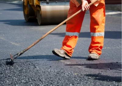 Worker in orange pants spreading asphalt with a rake, with a yellow road roller in the background.