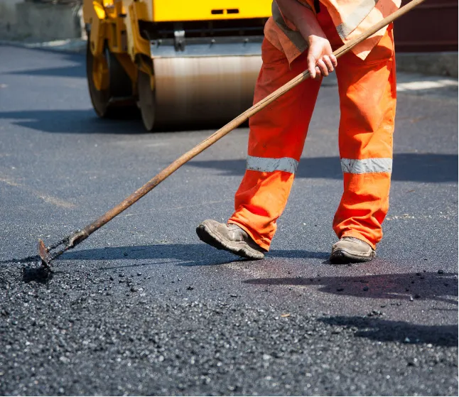 Construction worker in orange gear spreading asphalt on a road with a rake; roller machine in background.