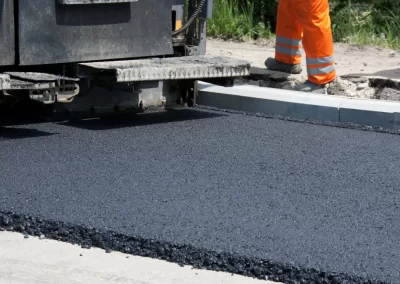 A paving machine lays fresh asphalt on a road as a worker in orange safety gear stands nearby.