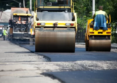 Two steamrollers flatten fresh asphalt on a road under construction, with workers and equipment visible.