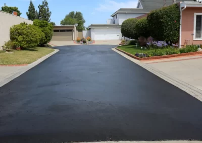 Freshly paved asphalt driveway between two houses, leading to garages, with greenery on both sides.