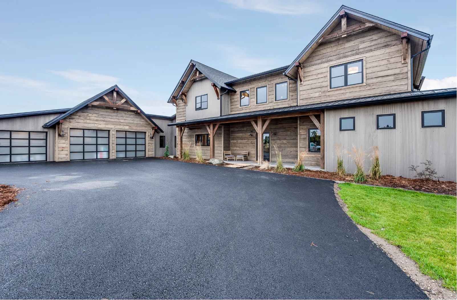 Modern rustic house with wood siding, large windows, attached garage, and paved driveway on a clear day.