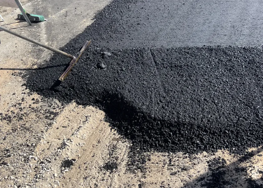 A rake spreads fresh asphalt over a road during paving work on a sunny day.