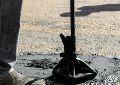 A person tamps down fresh asphalt on a road with a black tamping tool, wearing work boots.