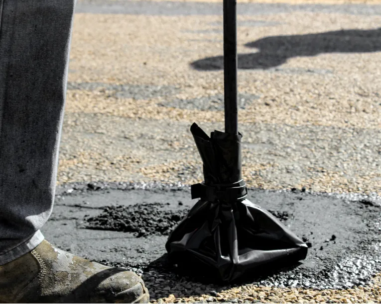 A person tamps down fresh asphalt on a road with a black tamping tool, wearing work boots.