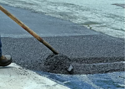 A person spreads fresh asphalt on a road with a shovel; an orange cone is visible in the background.