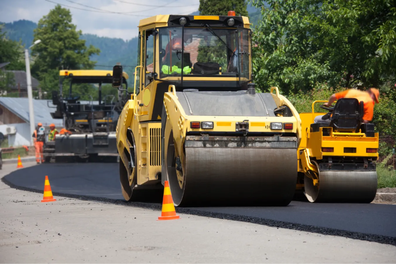 Road construction with yellow steamrollers flattening fresh asphalt, outlined by orange cones on a sunny day.