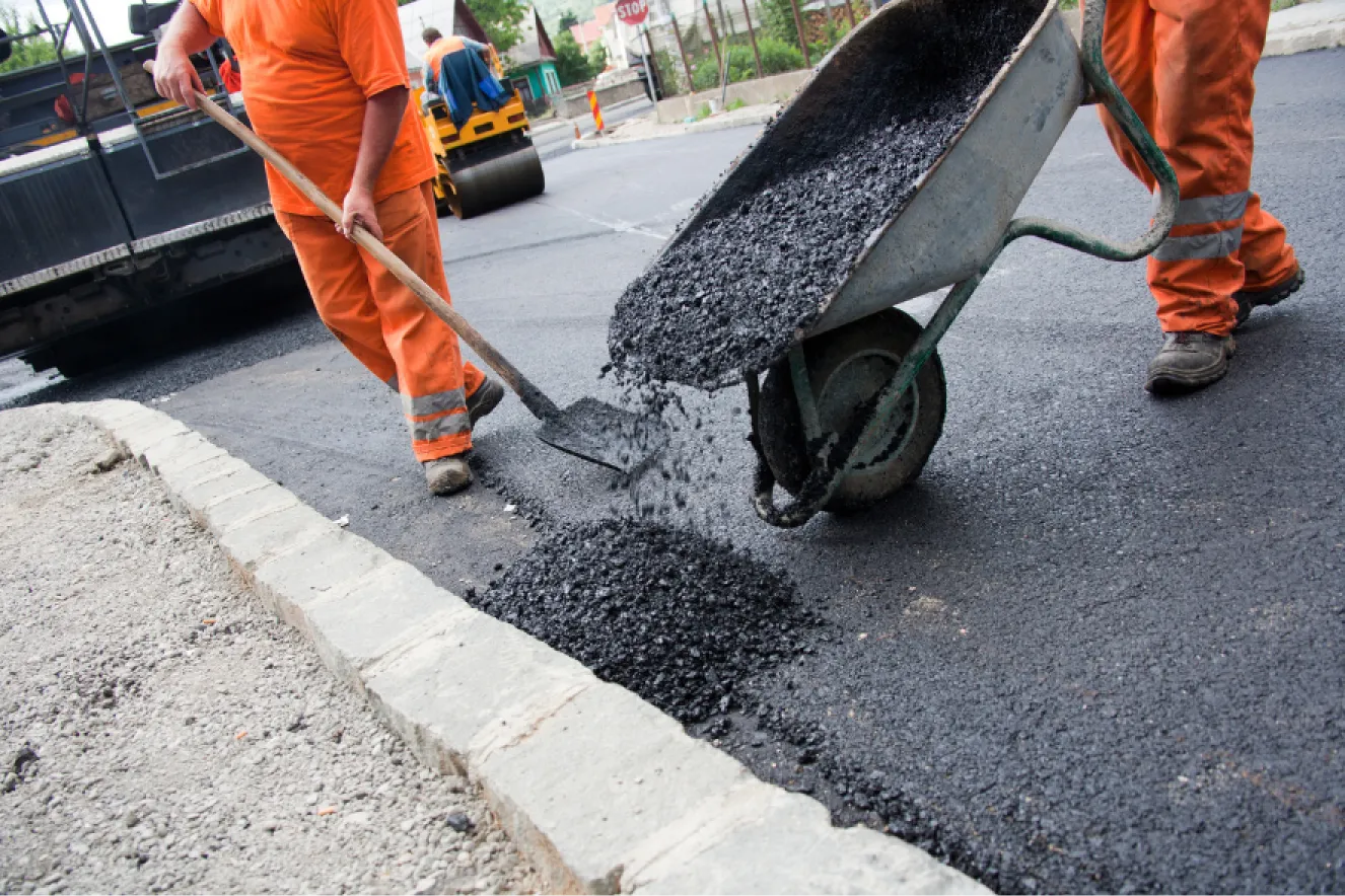 Workers in orange uniforms pour and spread asphalt from a wheelbarrow onto a road under construction.
