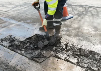 Worker in safety gear using a shovel to repair a road with broken asphalt and a traffic cone nearby.
