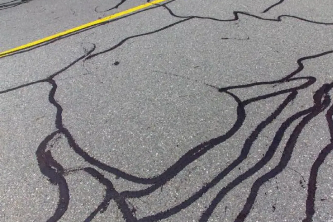 Cracks filled with black sealant form abstract lines and shapes on a gray asphalt road with a yellow divider line.