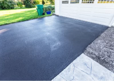 Freshly paved black asphalt driveway in front of a white garage, with a green lawn and trash bins nearby.