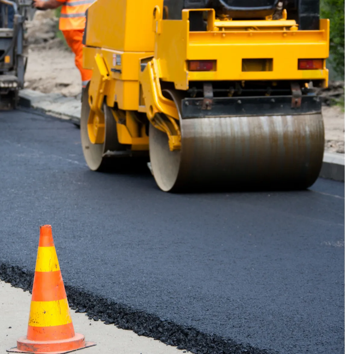 A road roller flattens fresh asphalt with an orange traffic cone in the foreground.