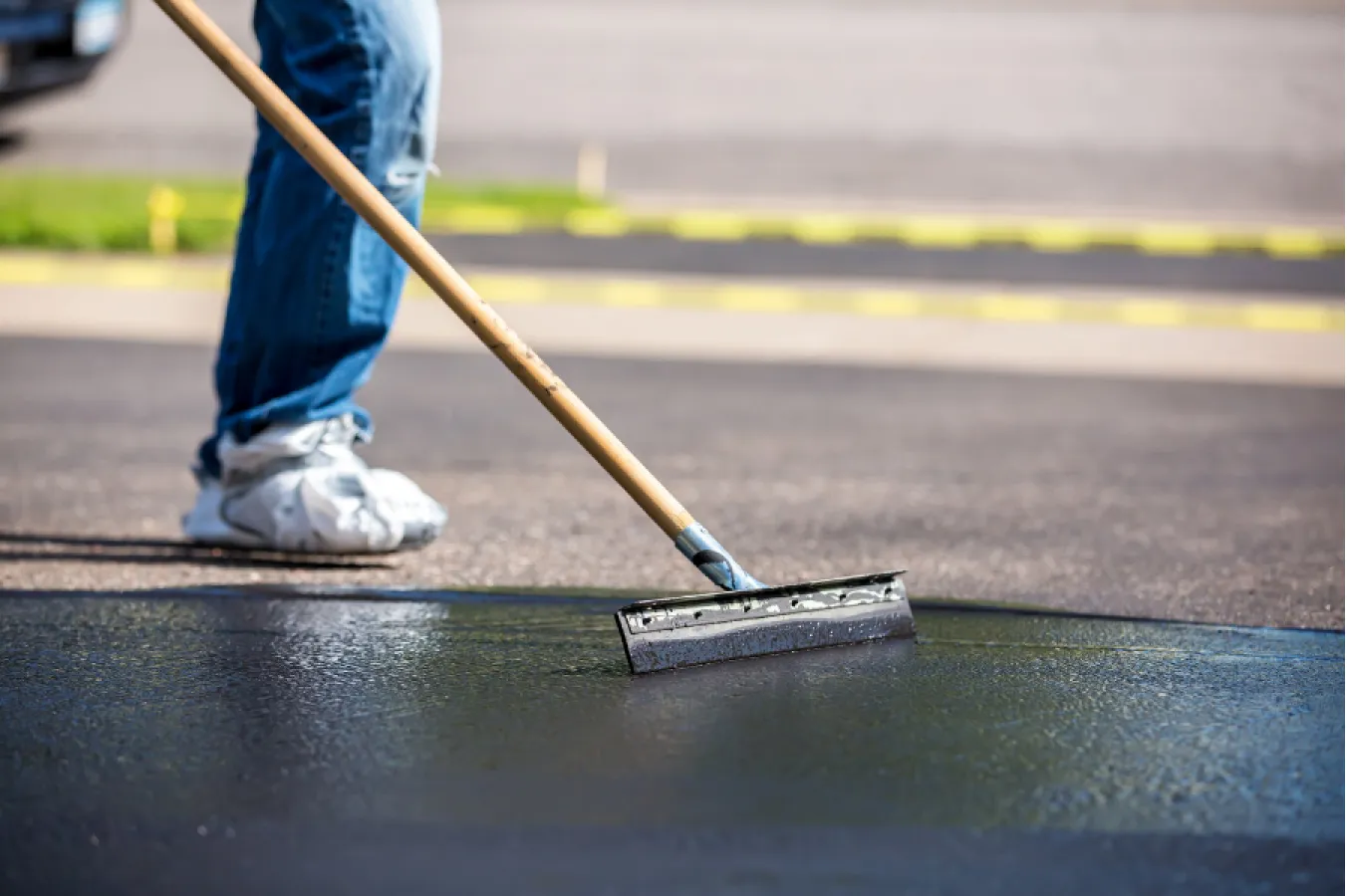 Person spreading black sealant on asphalt with a squeegee tool; only legs and tool are visible.