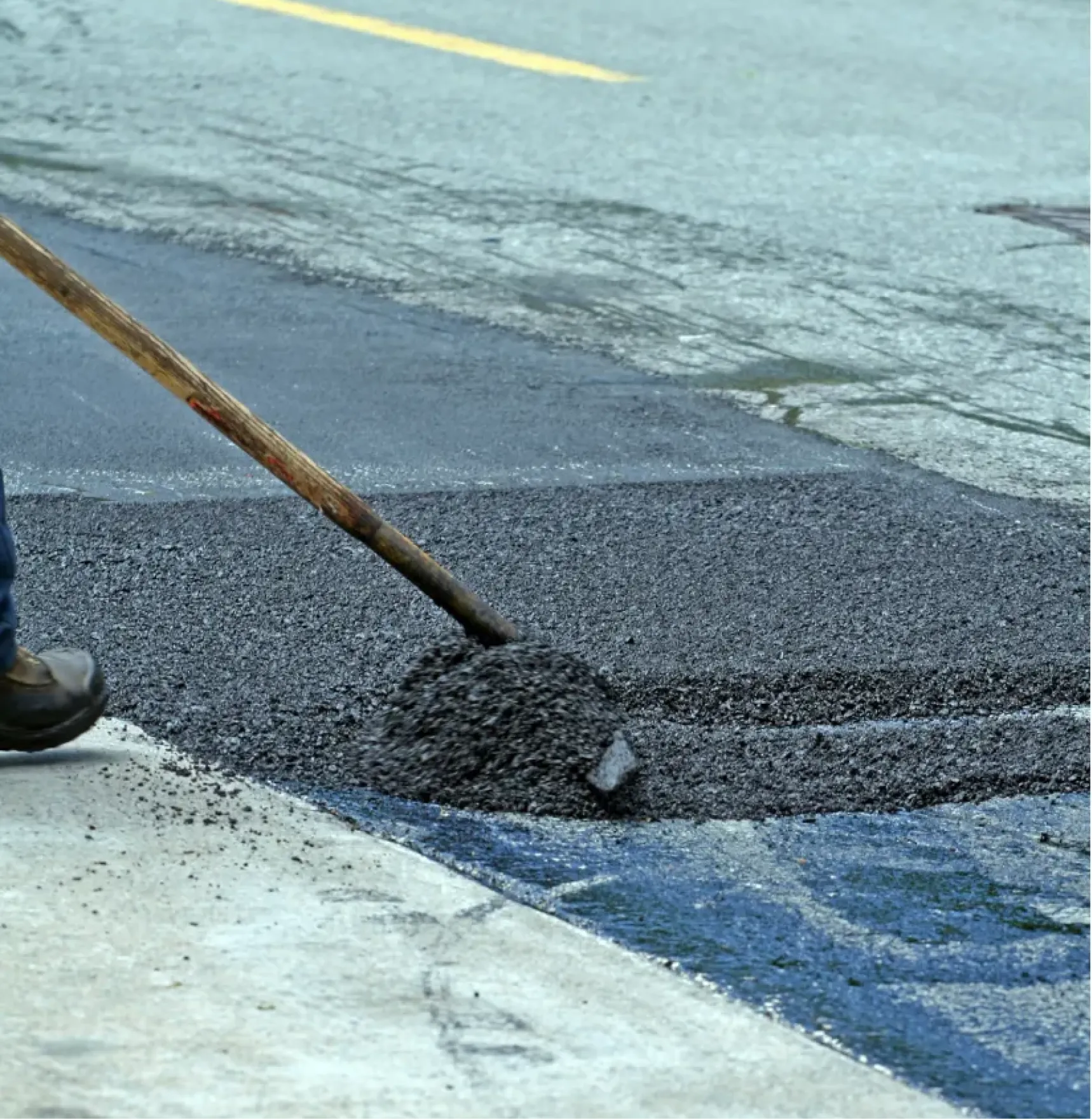 A worker spreads fresh asphalt on a cracked road using a shovel.