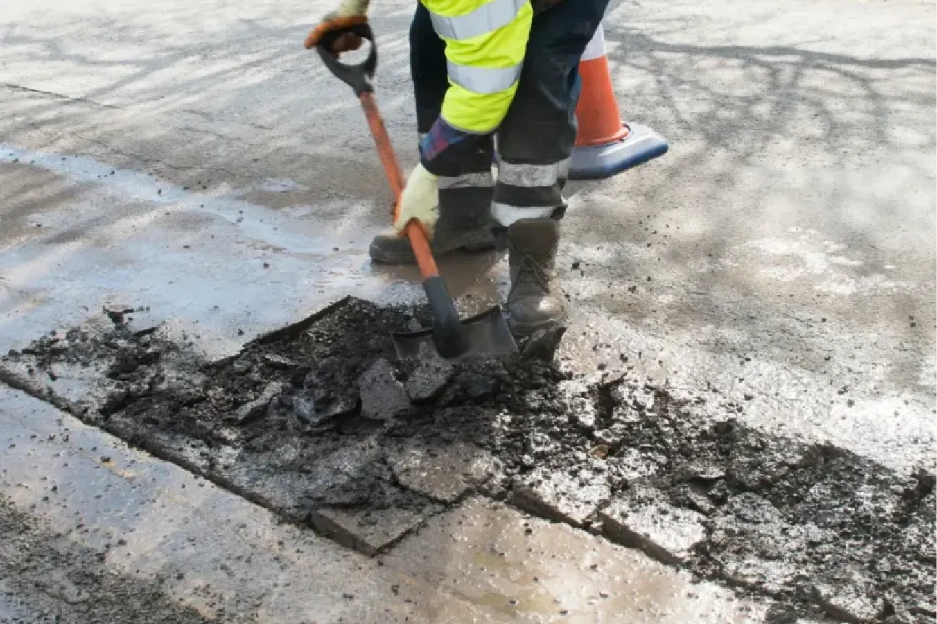 Worker in reflective gear using a shovel to repair a damaged asphalt road near an orange traffic cone.