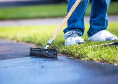 Person sealing a driveway with a long-handled tool, wearing jeans and shoe covers, near green grass.