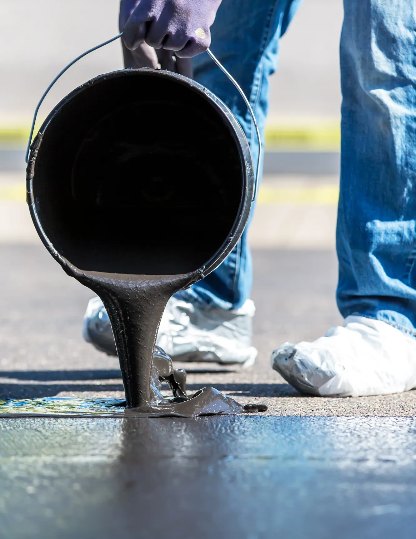 Person pouring black tar from a bucket onto pavement, wearing gloves and shoe covers, outdoors.