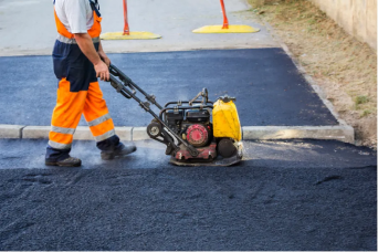 Worker operating a plate compactor on freshly laid asphalt pavement near a curb.